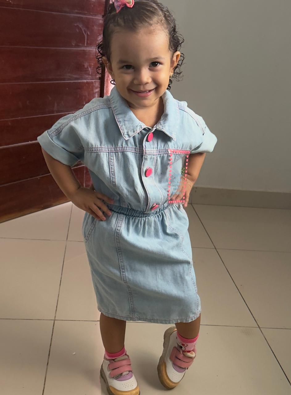 Milena sorrindo, com as mãos na cintura, vestido jeans e tênis com detalhes cor-de-rosa.
